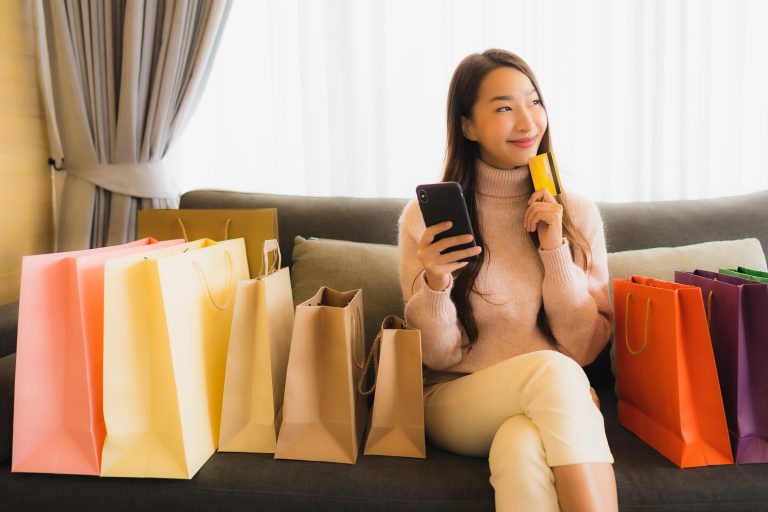 Portrait beautiful young asian woman using laptop or smart mobile cell phone for shopping online on sofa around shopping bag in living room interior